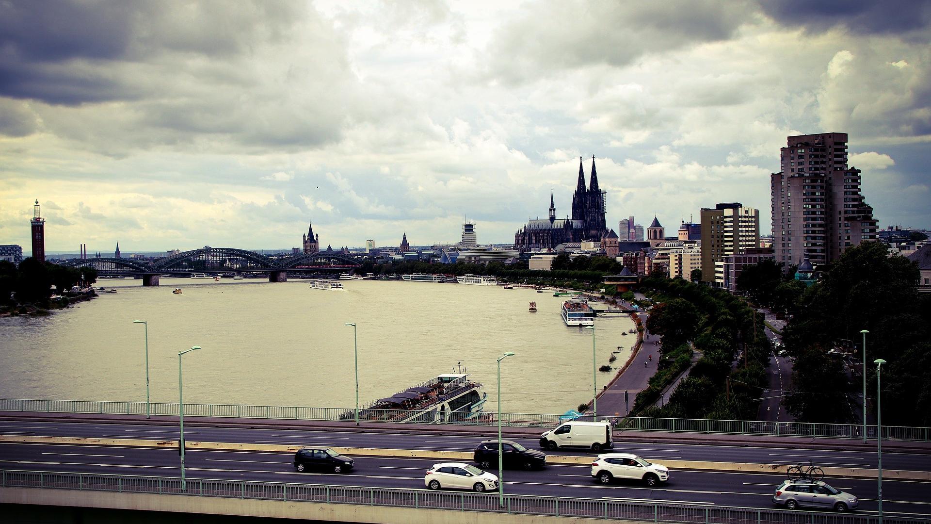 Blick auf Köln mit dem Rhein und dem Kölner Dom im Hintergrund. Eine Brücke und Autos im Vordergrund, dramatischer Himmel.