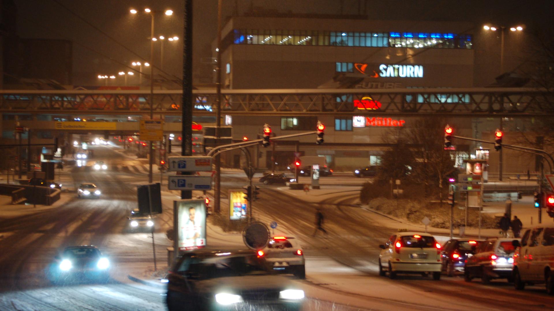 Winterliche Nacht in Wuppertal: Eine verschneite Straße in Wuppertal, beleuchtet von den Lichtern vorbeifahrender Autos und markanten Gebäudeschildern wie Saturn und Müller, erzeugt eine stimmungsvolle urbane Atmosphäre.