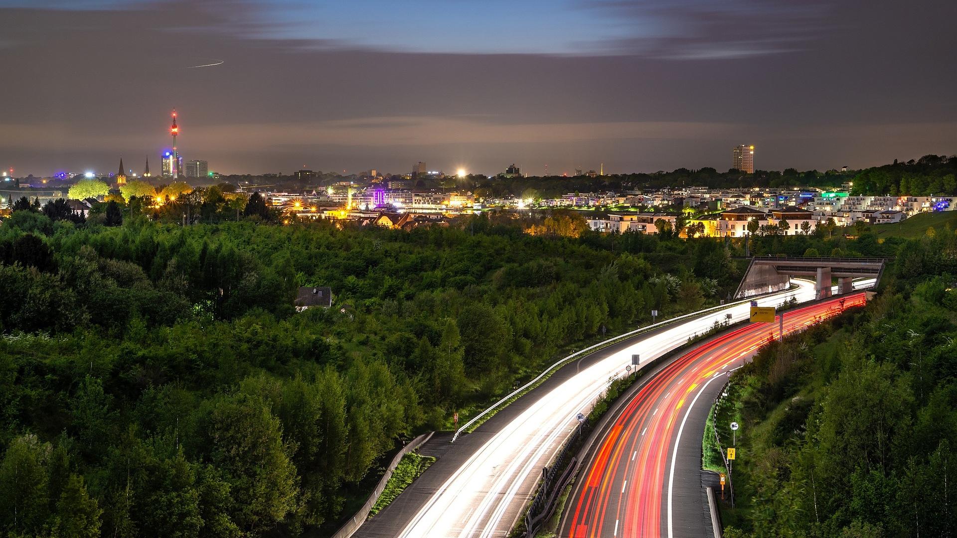 Das Bild zeigt die nächtliche Skyline von Dortmund, Nordrhein-Westfalen, mit leuchtenden Stadtlichtern und einer stark befahrenen Autobahn im Vordergrund. Die langen Lichtspuren der Autos vermitteln Dynamik und Bewegung, während die üppige Vegetation den Kontrast zur urbanen Umgebung betont. Eine malerische Aussicht auf die Stadt, die das pulsierende Nachtleben und die Verkehrsanbindung von Dortmund in Szene setzt.