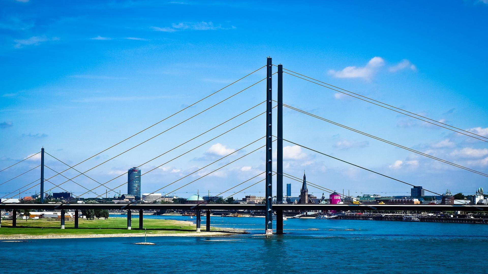 Dieses Bild zeigt die Rheinkniebrücke in Düsseldorf, eine elegante Schrägseilbrücke über den Rhein. Im Hintergrund erstreckt sich die Skyline der Stadt mit modernen Gebäuden und historischen Bauwerken. Die Szene hebt die Verbindung von Architektur und Natur unter einem klaren blauen Himmel hervor.