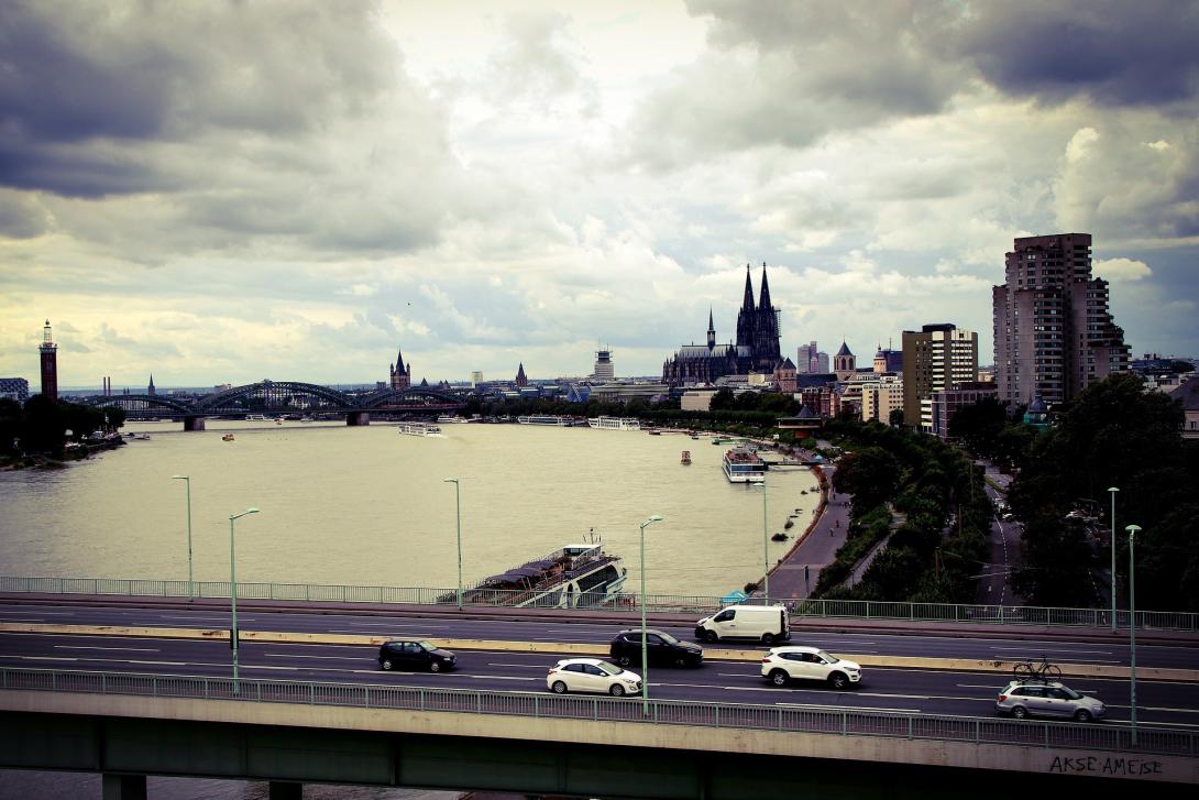 Blick auf Köln mit dem Rhein und dem Kölner Dom im Hintergrund. Eine Brücke und Autos im Vordergrund, dramatischer Himmel.