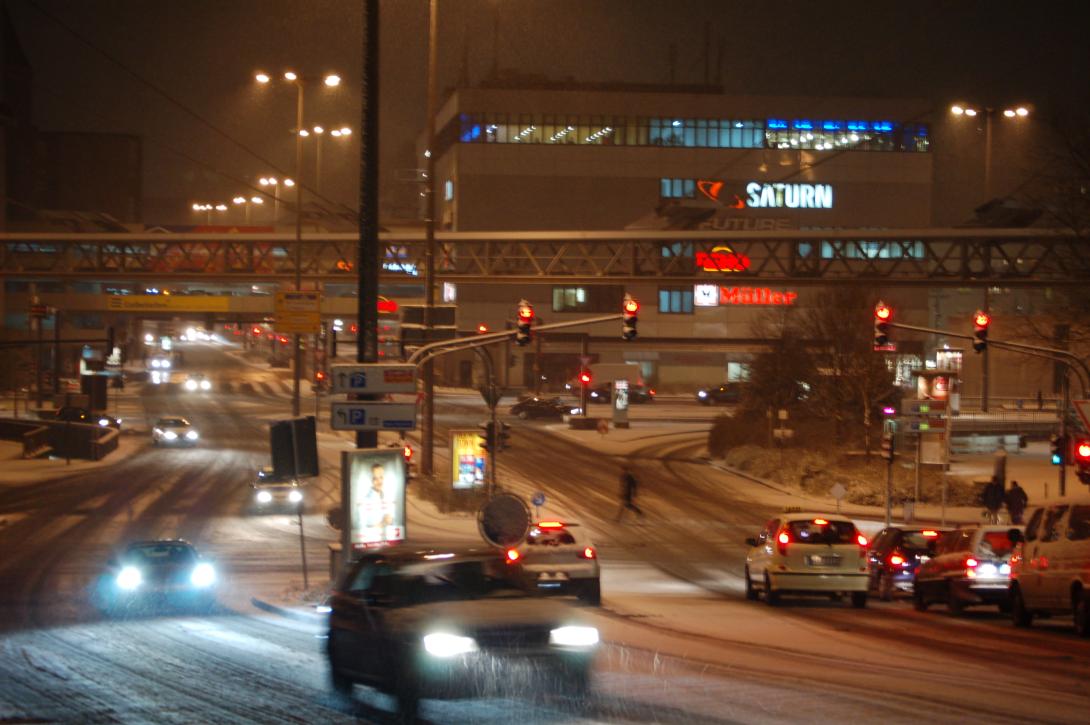 Winterliche Nacht in Wuppertal: Eine verschneite Straße in Wuppertal, beleuchtet von den Lichtern vorbeifahrender Autos und markanten Gebäudeschildern wie Saturn und Müller, erzeugt eine stimmungsvolle urbane Atmosphäre.