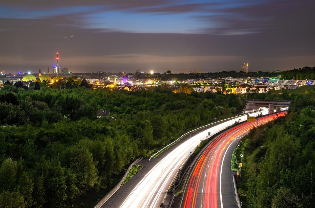 Das Bild zeigt die nächtliche Skyline von Dortmund, Nordrhein-Westfalen, mit leuchtenden Stadtlichtern und einer stark befahrenen Autobahn im Vordergrund. Die langen Lichtspuren der Autos vermitteln Dynamik und Bewegung, während die üppige Vegetation den Kontrast zur urbanen Umgebung betont. Eine malerische Aussicht auf die Stadt, die das pulsierende Nachtleben und die Verkehrsanbindung von Dortmund in Szene setzt.