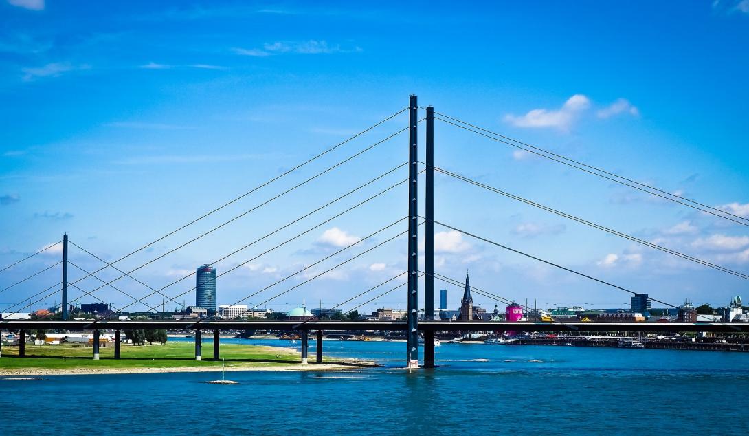 Dieses Bild zeigt die Rheinkniebrücke in Düsseldorf, eine elegante Schrägseilbrücke über den Rhein. Im Hintergrund erstreckt sich die Skyline der Stadt mit modernen Gebäuden und historischen Bauwerken. Die Szene hebt die Verbindung von Architektur und Natur unter einem klaren blauen Himmel hervor.